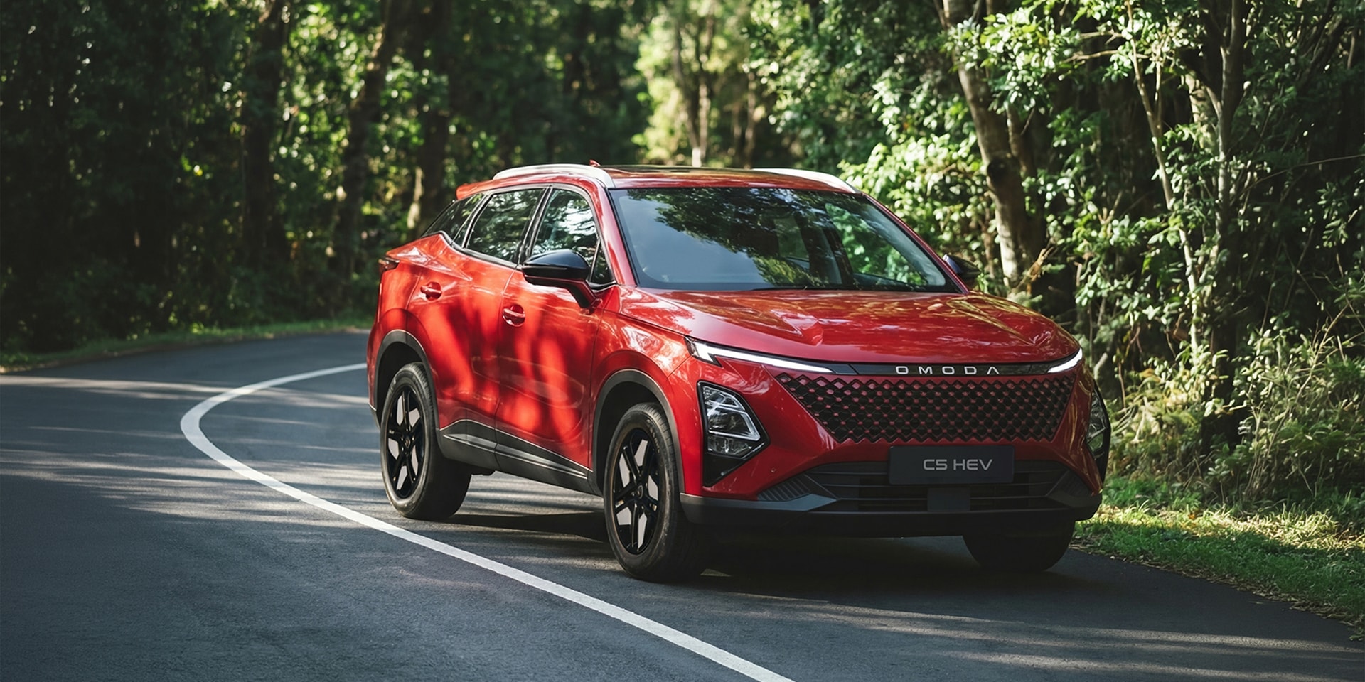 Red Omoda CS HEV SUV driving on a curving forest road with sunlight filtering through trees.